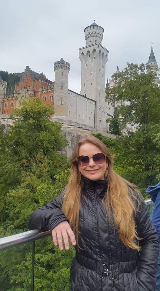 Mulher branca loira de cabelos longos em frente ao Castelo de Newschwanstein, em Füssen na Alemanha. Nas laterais da foto tem árvores típicas de regiões mais frias. A mulher, Eliete, usa óculos escuros e um casaco preto de frio.