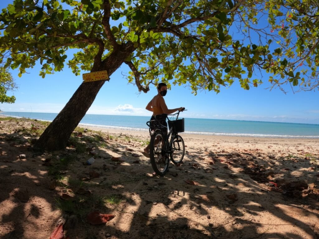 Mulher na praia de máscara com uma bicicleta abaixo da sombra de uma árvore
