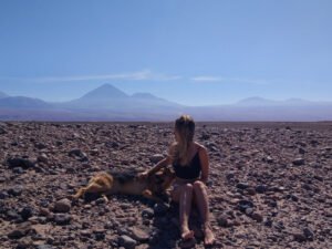 Mulher no deserto do Atacama com um cachorro ao lado. De fundo, dá para ver 2 vulcões.