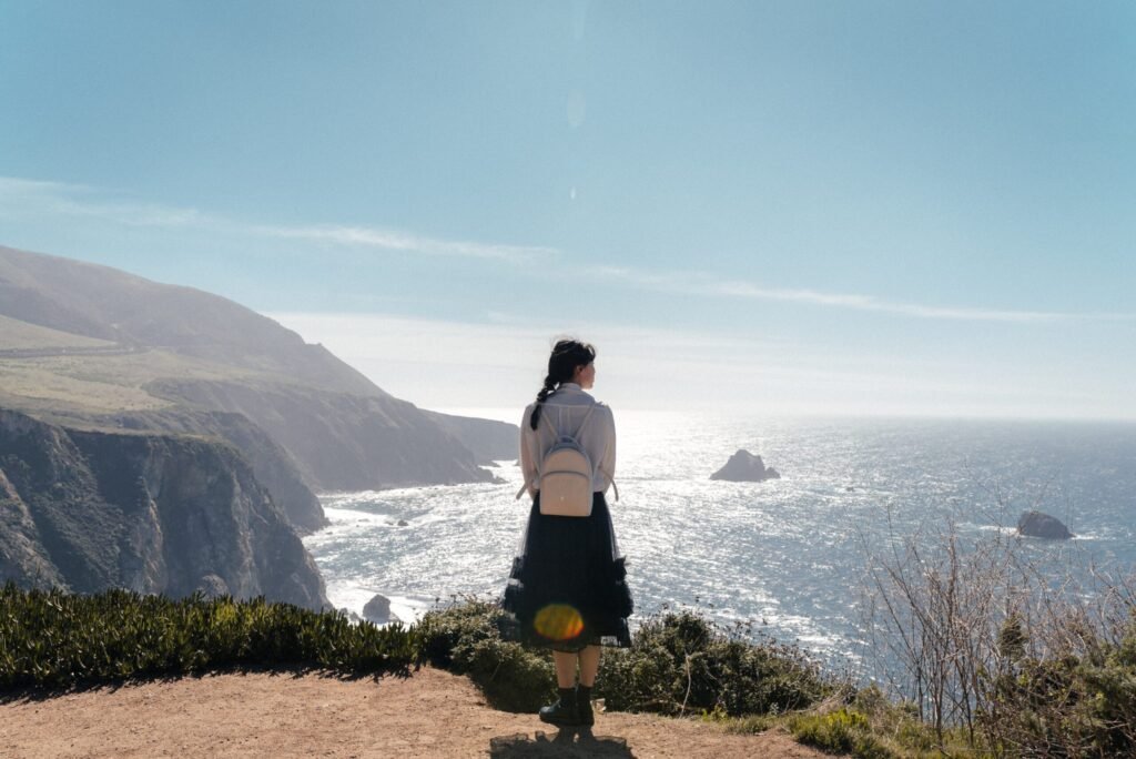 Mulher com trança preta de costas com uma mochila admirando uma paisagem de falésias e o oceano refletindo a luz do sol.