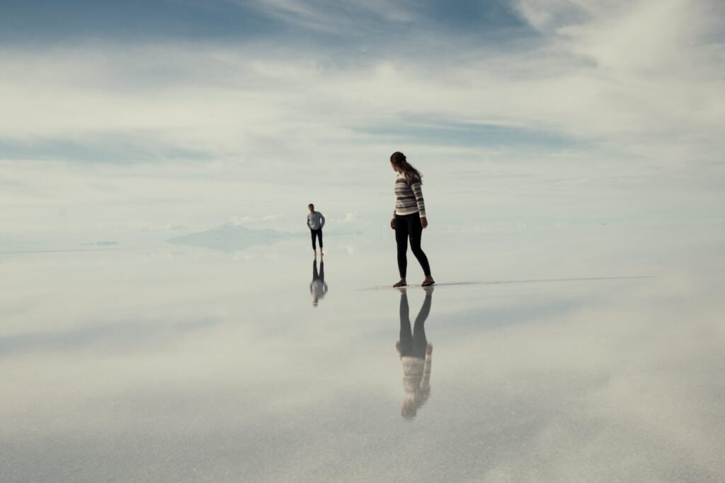 Foto do Salar do Uyuni, deserto de sal, com água no chão espelhando o céu e duas pessoas que andam sob a água.+