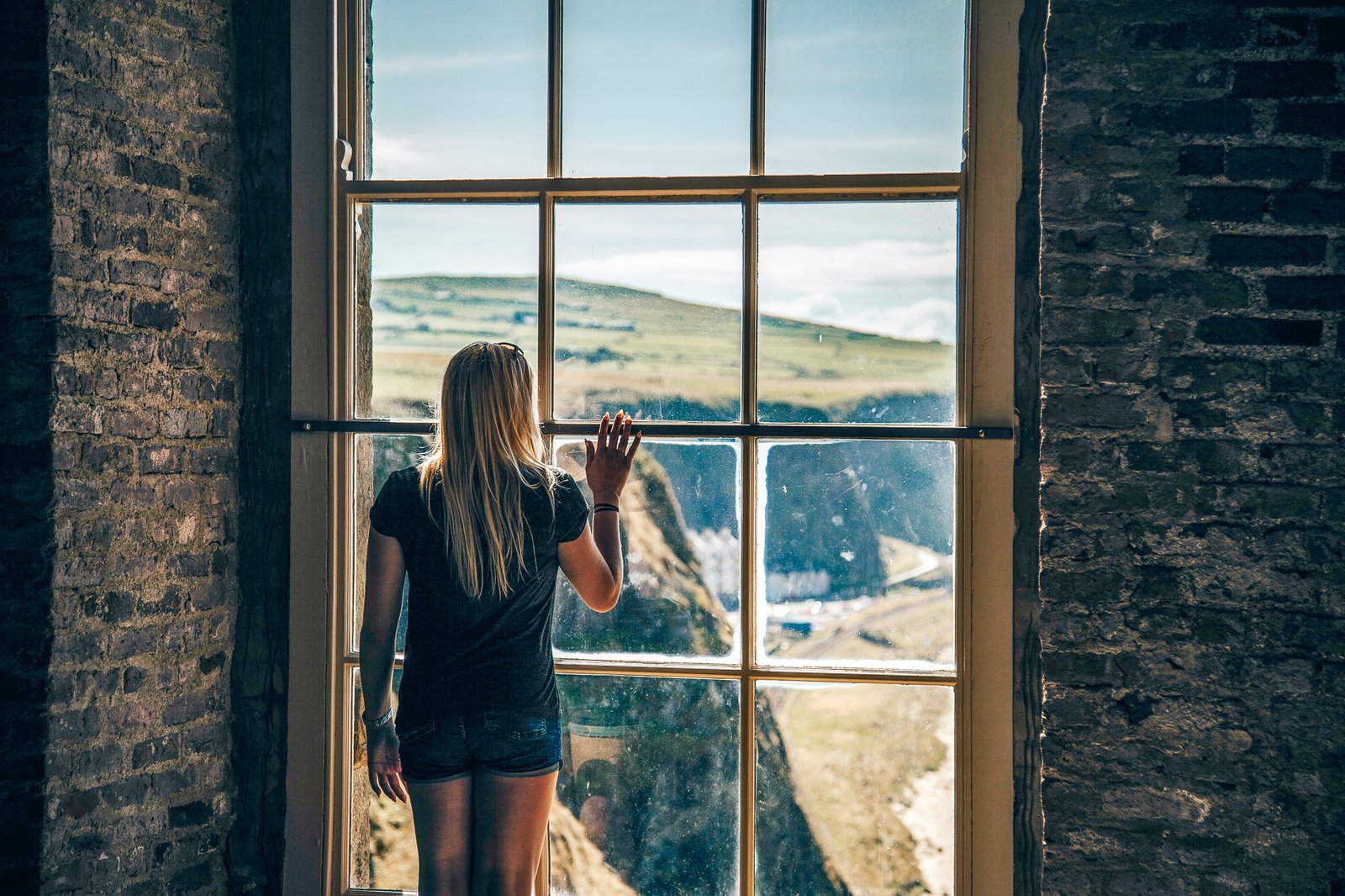 Mulher loira de costas pra uma janela com paisagem de montanhas a frente.